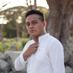 A confident man standing outdoors in a white shirt, captured in soft natural light.