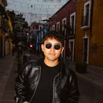A stylish young man wearing sunglasses and a leather jacket on a colorful urban street.